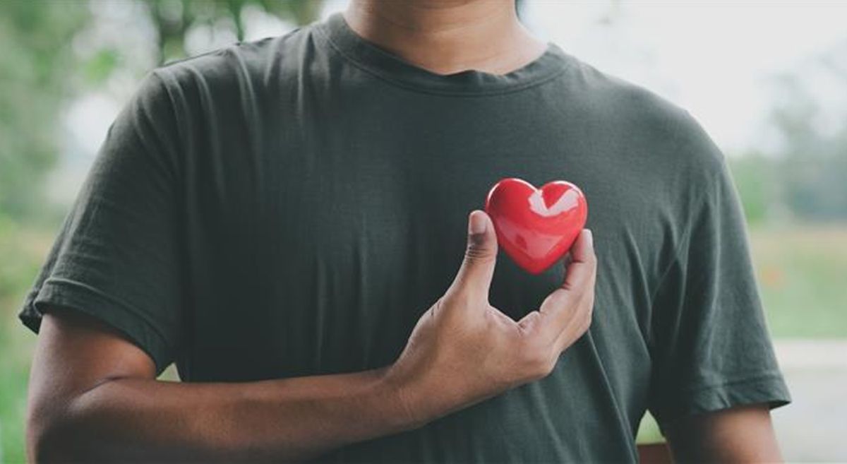 Male wearing a dark teeshirt and holding a red heart shaped object at his heart.
